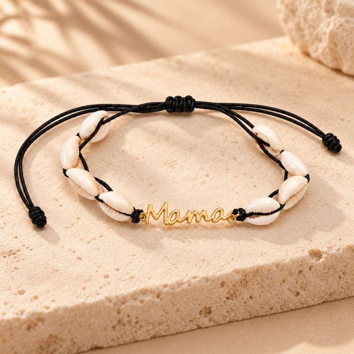 Pulsera Mamá con conchas blancas, cordón negro ajustable y dije dorado sobre fondo cálido